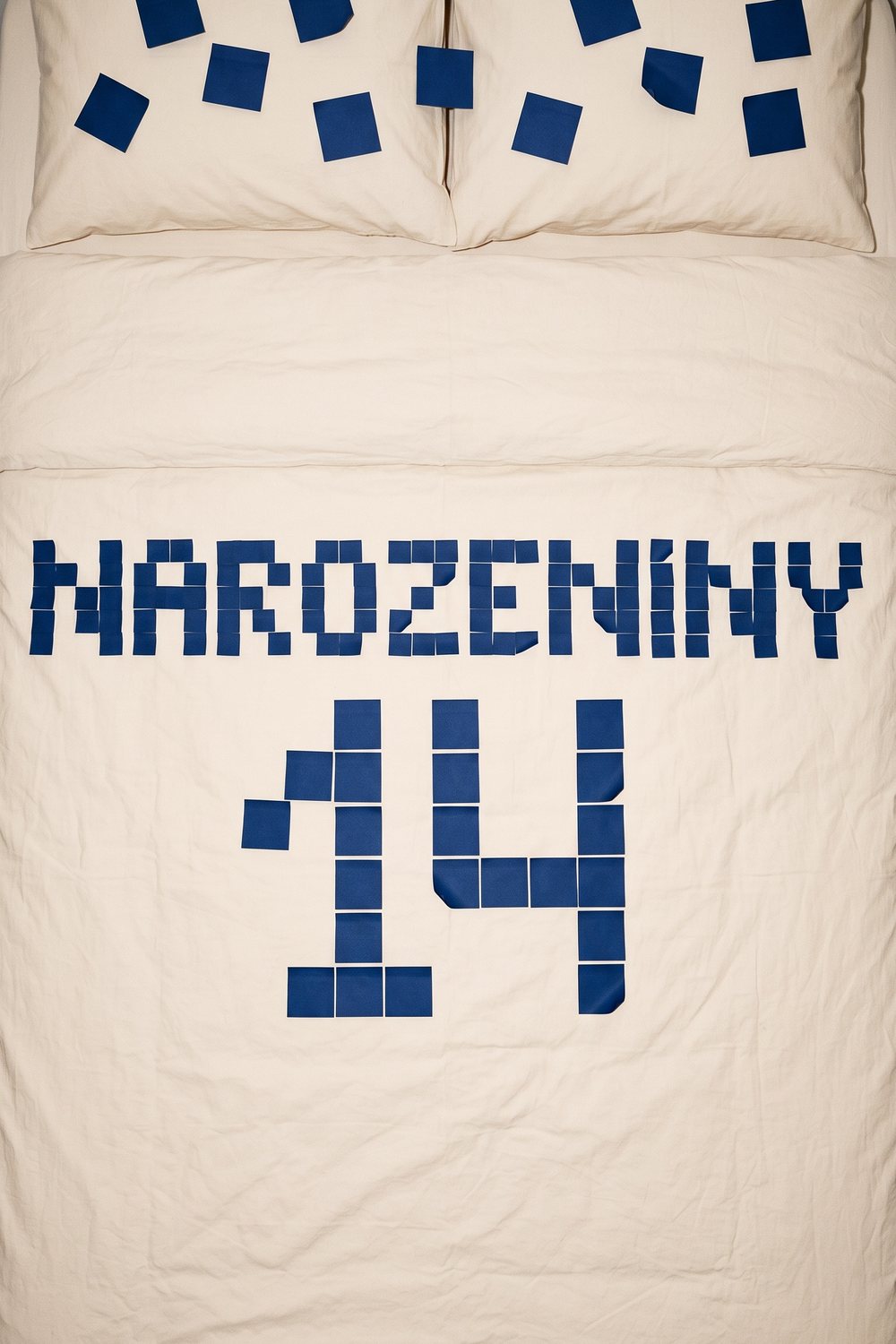 Ustlaná postel, na přikrývce je z modrých čtverečků poskládaný nápis NAROZENINY 14